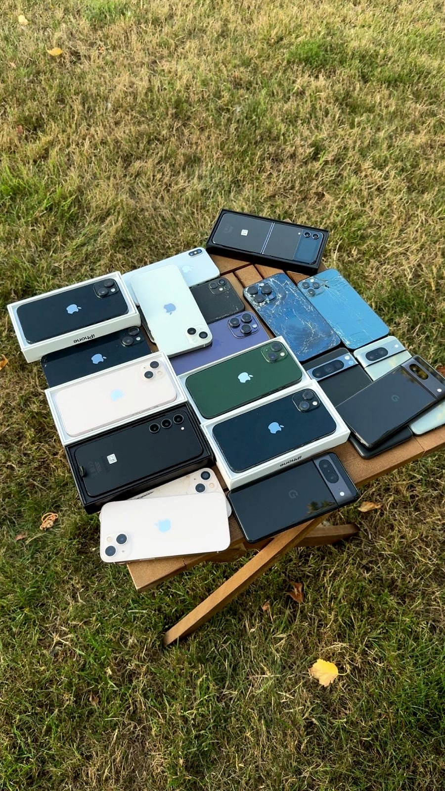 A batch of phones laid out on a garden table, some with cracked backs