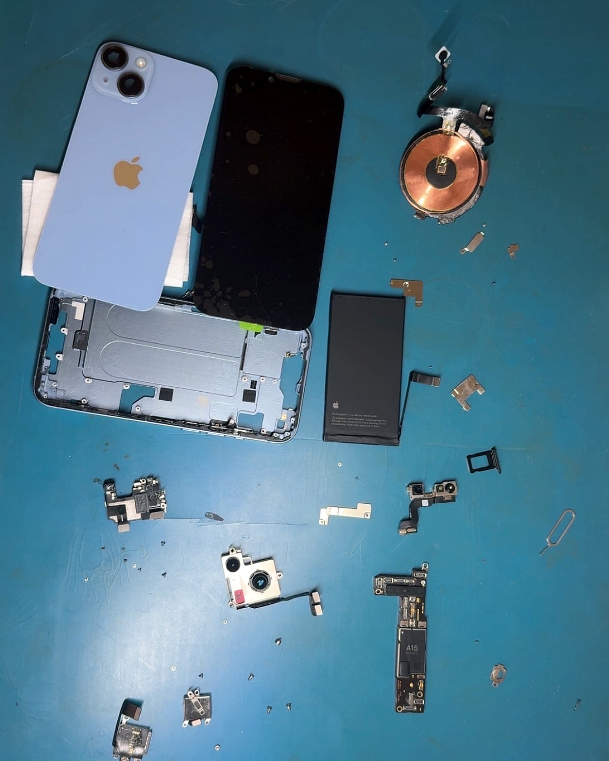An iPhone 14 taken apart on the bench: back glass, screen, frame, battery, charging coil, cameras and logic board laid out